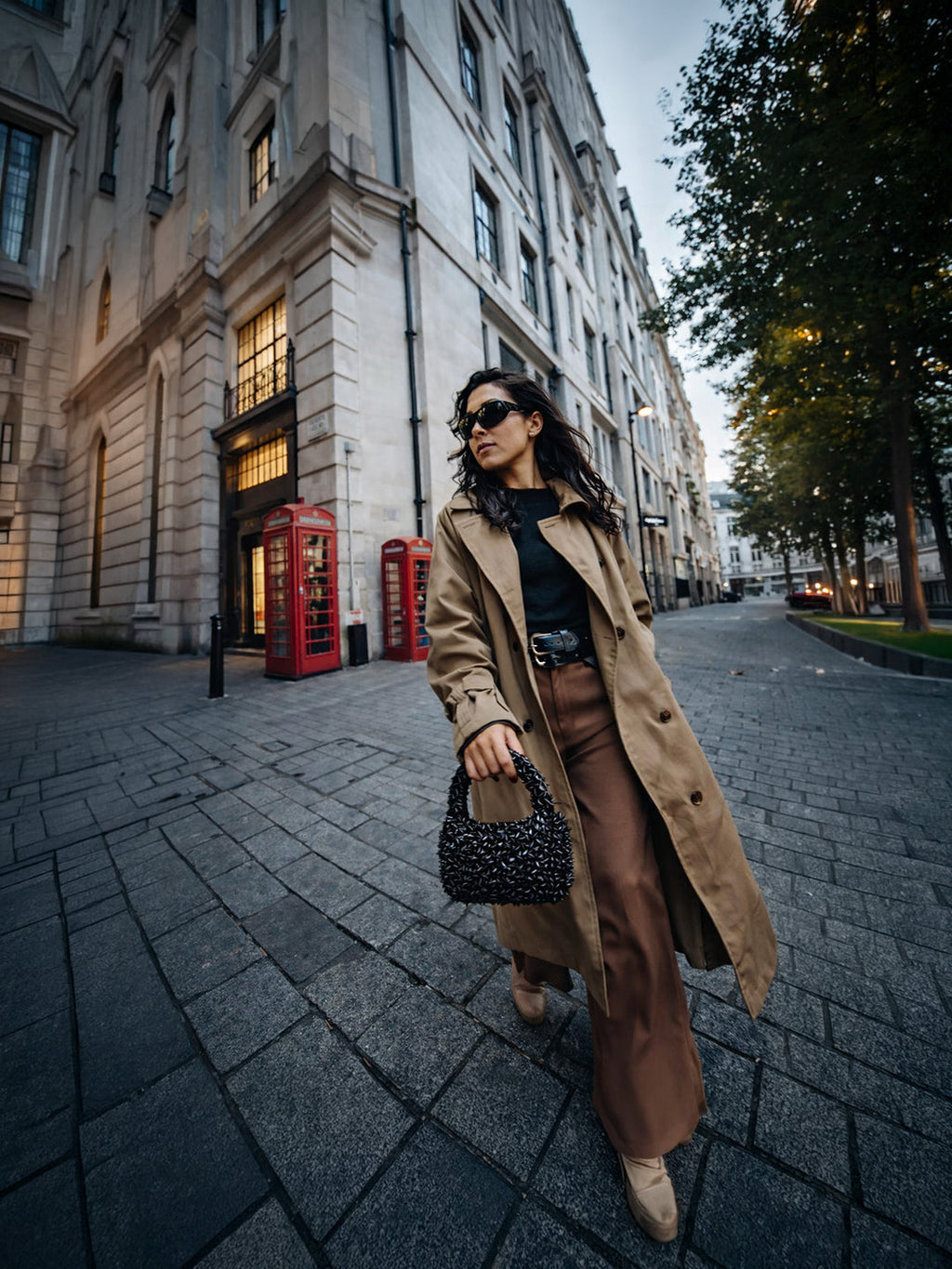 Woman walking down a city street wearing a beige trench coat and sunglasses.