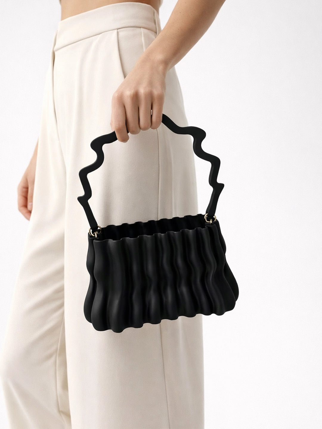Black ruffled handbag held by a person wearing white pants on a white background